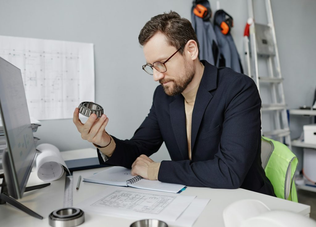 Bearded mechanical engineer holding machine part at workplace